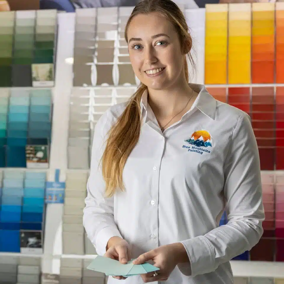 A woman in a white shirt holds paint swatches in a store with a wall display of colorful paint samples behind her, meticulously planning her next interior design project inspired by the serene hues of the Blue Mountains.