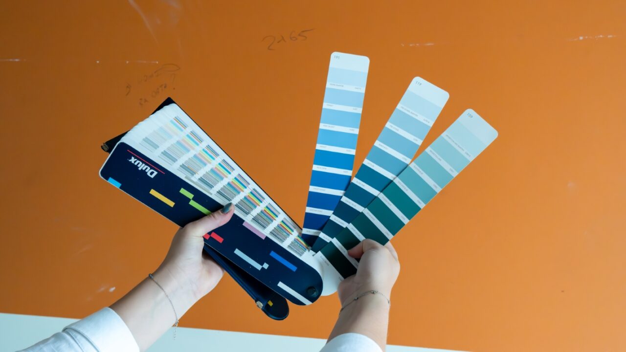 Person holding paint swatches against an orange wall, comparing various shades of blue and green.
