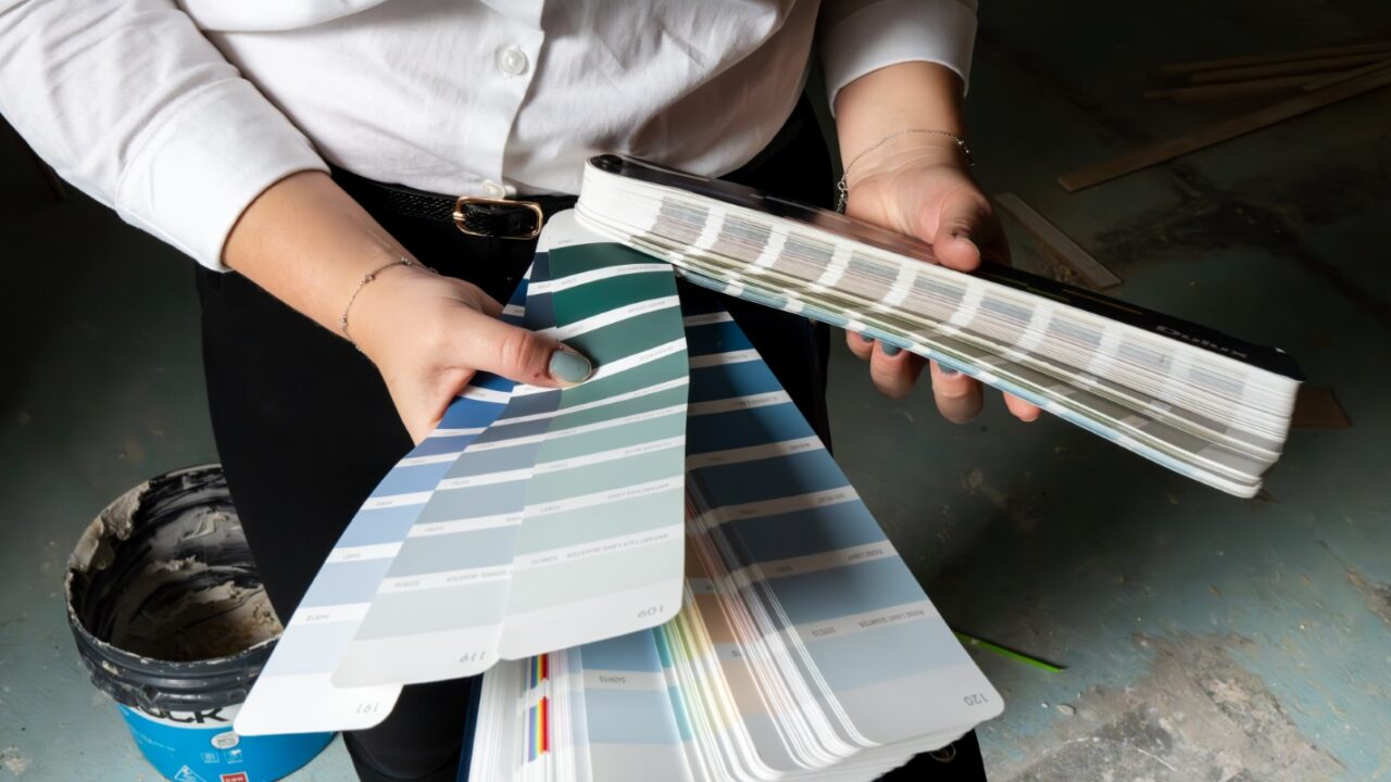 A person holding a spread of paint color swatches in both hands, standing near a bucket on a textured floor.