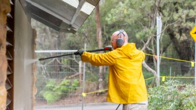 Person in a yellow raincoat pressure washing a building's exterior, surrounded by trees and caution tape.