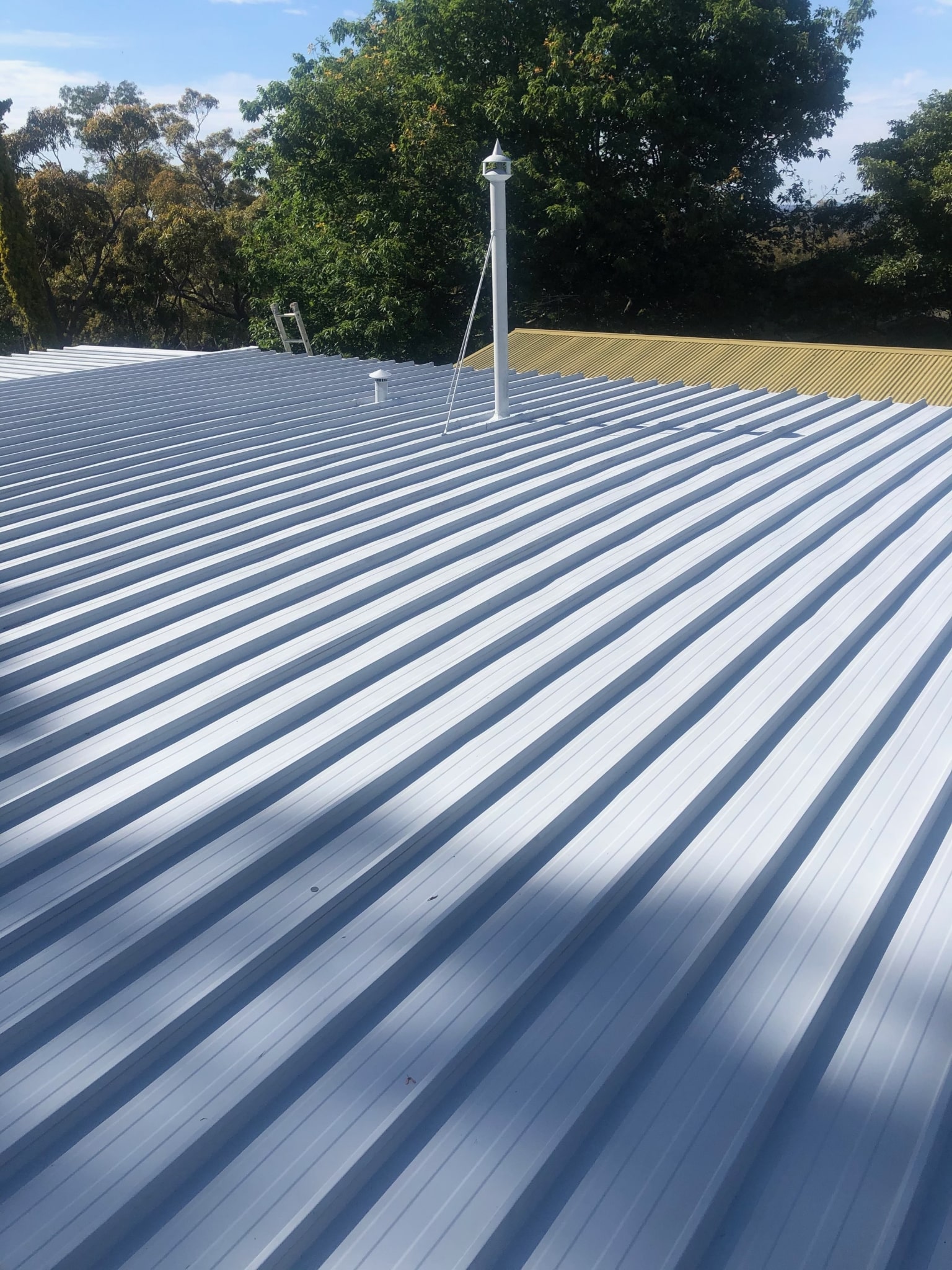 A corrugated metal roof with a vent pipe under a clear blue sky, surrounded by trees