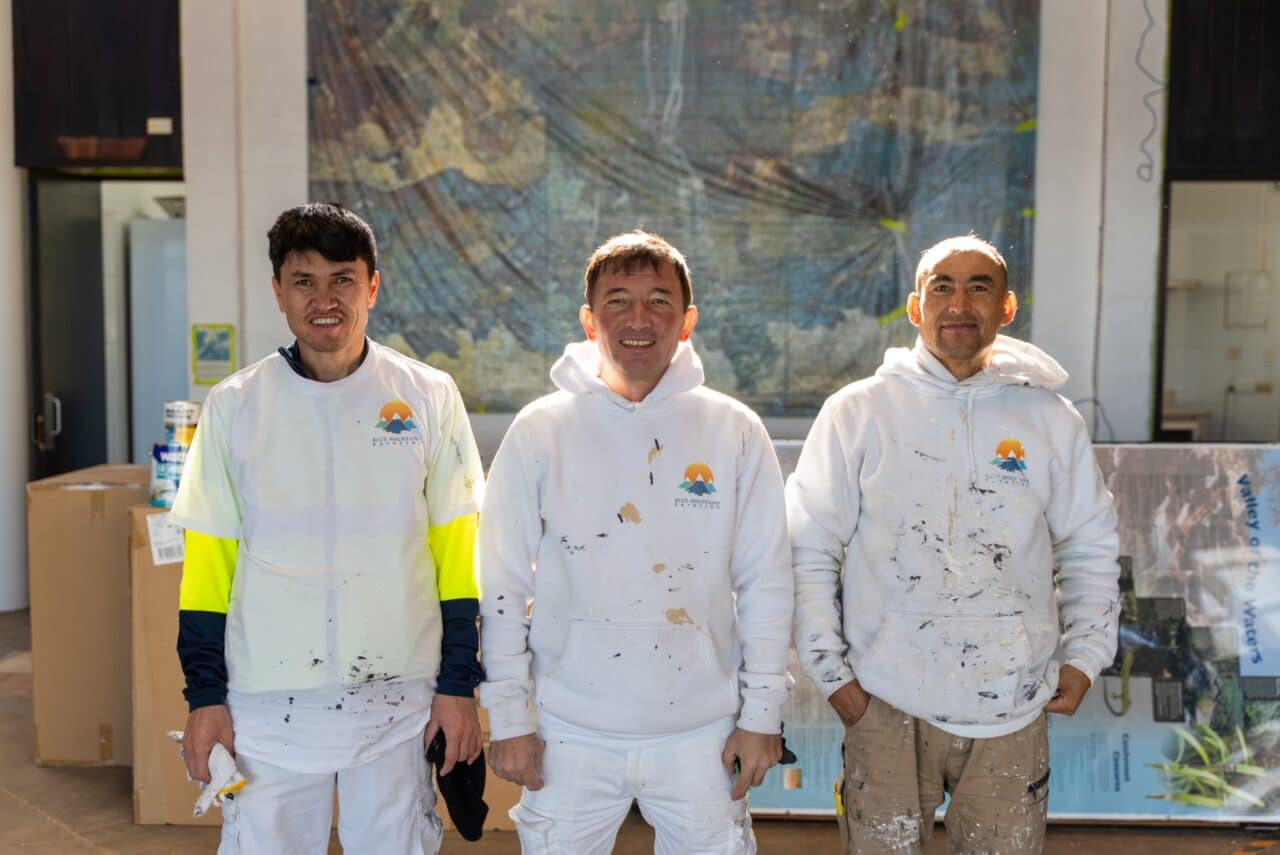 Three painters in paint-spattered clothing stand in front of a mural, smiling at the camera.