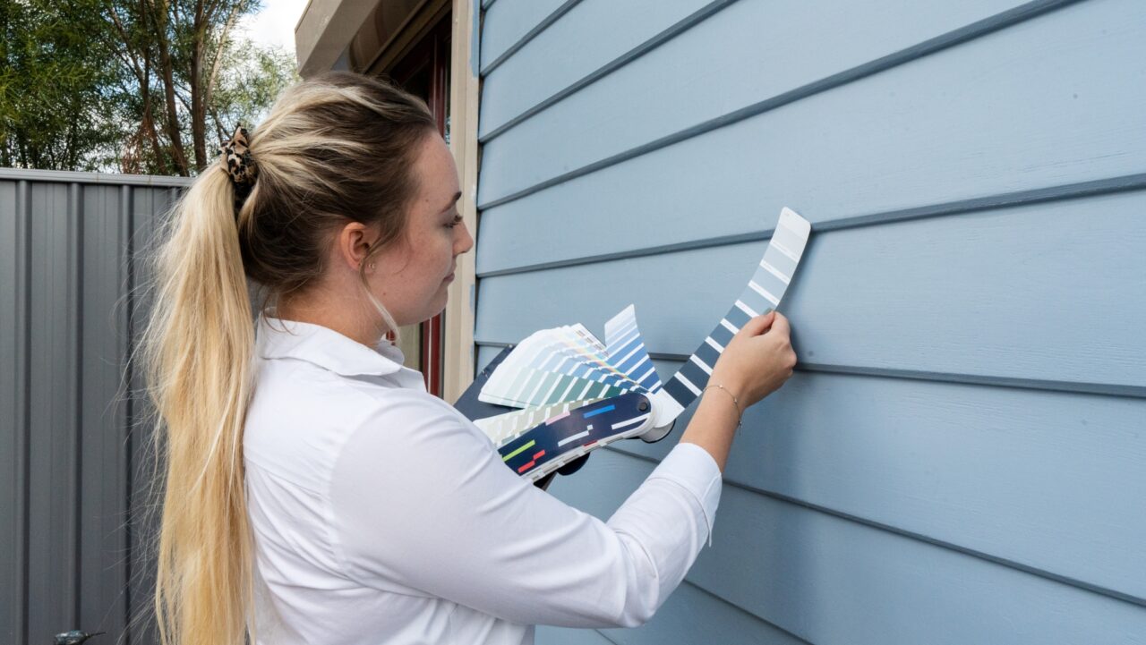 A woman with long blonde hair holds up paint swatches against blue exterior house