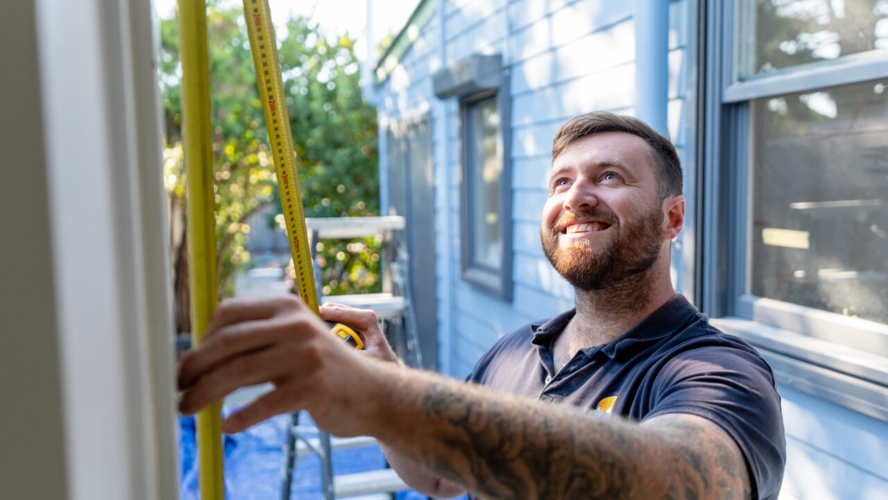 Man in polo shirt measuring wall outdoors with tape, in front of light blue house