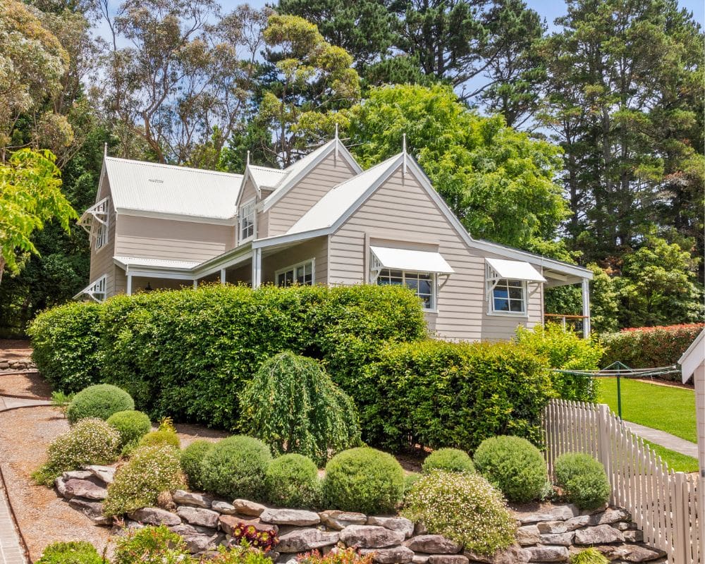 a two-story beige house with white trim and a metal roof, surrounded by green bushes