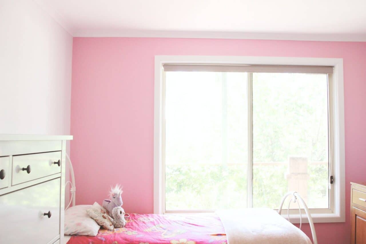 a pink and white bedroom with a bed, stuffed animals, a dresser, and a large window letting in natural light showcases timeless interior paint inspired by the latest interior painting trends.