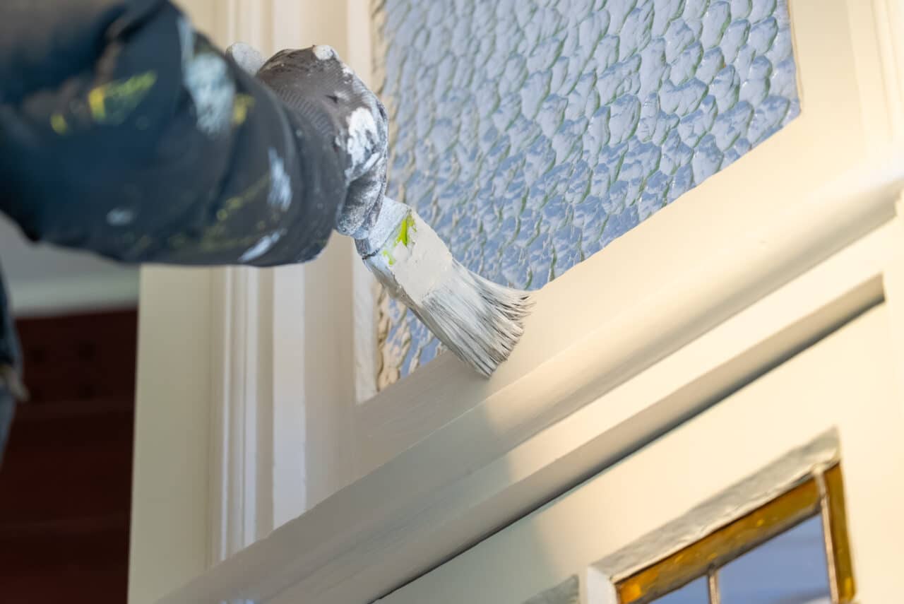 the hand of a painter painting white paint on a door with a window